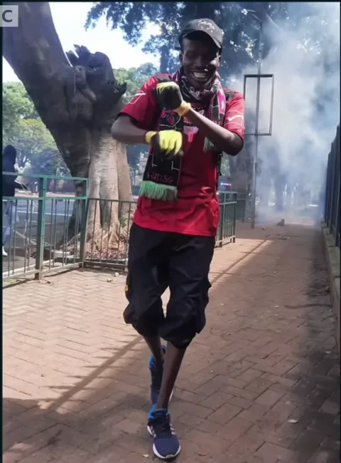 Eric Shieni on the front line in a red top, black shorts and yellow gloves, tear gas behind him.